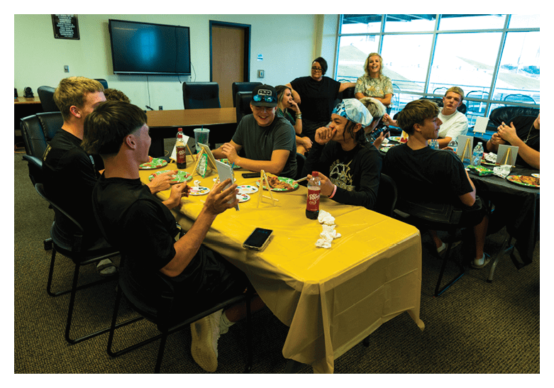 A group of people sitting around a table covered with a yellow tablecloth are eating pizza and engaging in conversation in a brightly lit room with large windows. Description generated by AI