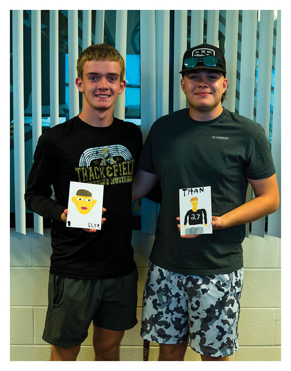 Two adults stand side by side indoors holding small drawings, one wearing a black long-sleeve shirt and shorts and the other wearing a dark t-shirt, patterned shorts, and a cap. Description generated by AI