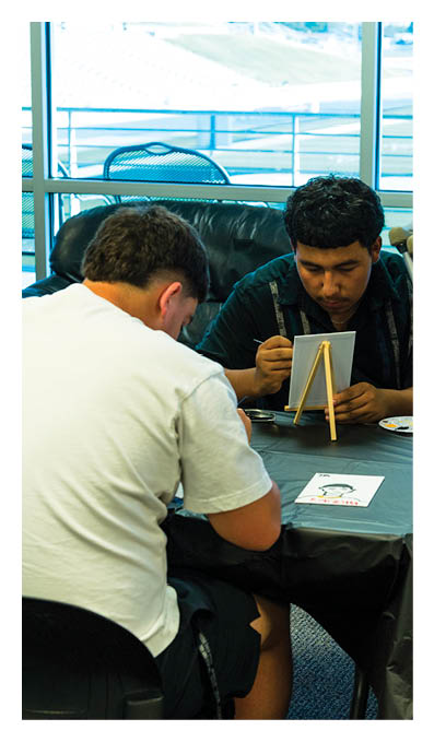 Two people sit at a table indoors with one painting on a small easel and the other drawing on paper, with crutches leaning against the table. Description generated by AI
