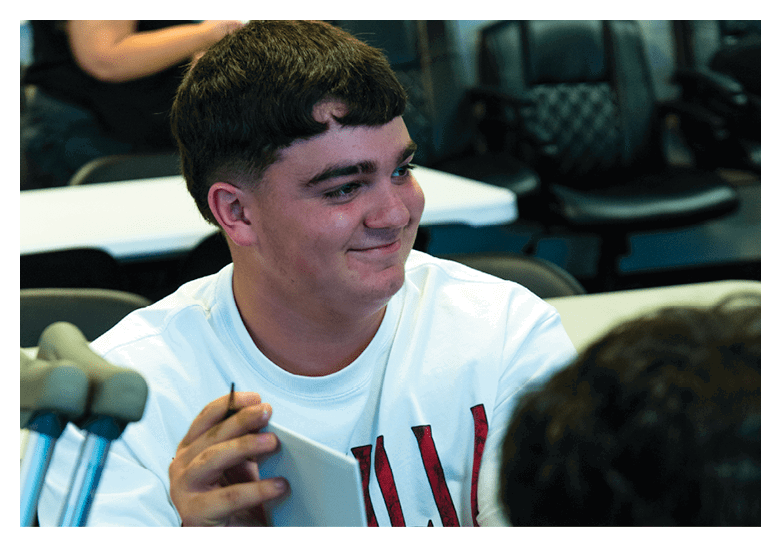 A person with short dark hair wearing a white shirt is smiling and looking to the side while sitting indoors with chairs and another person blurred in the background. Description generated by AI