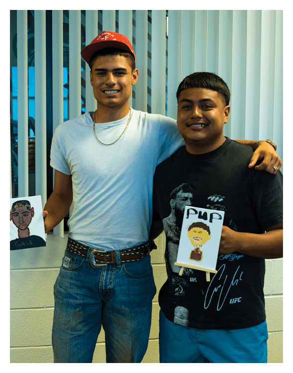 Two adults stand side by side indoors smiling, each holding a small canvas with a painted portrait, one wearing a red cap and white shirt and the other in a black graphic t-shirt and blue shorts. Description generated by AI
