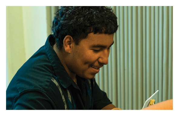 A person with short curly hair wearing a dark jacket is smiling and looking downward against a background of vertical blinds. Description generated by AI