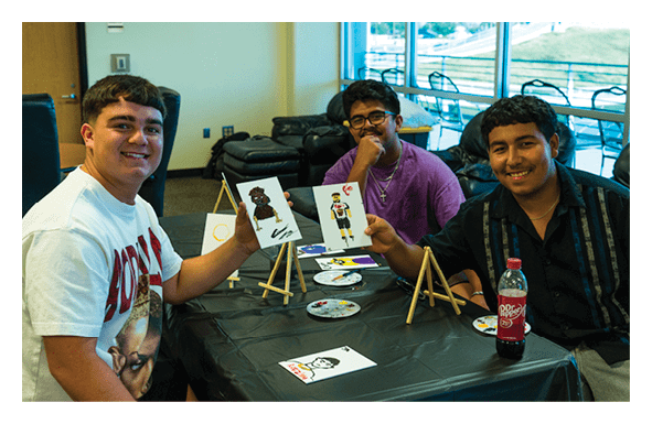 Three adults sitting at a table covered with a black cloth are smiling and holding up small paintings while engaging in a painting activity in a well-lit room. Description generated by AI