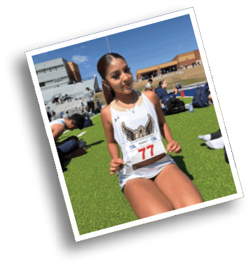 Person wearing a white athletic jersey with a winged logo and holding a race bib numbered 77 at an outdoor track and field event. Description generated by AI