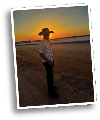 A person wearing a cowboy hat and a striped shirt stands on a beach at sunset with footprints visible in the sand. Description generated by AI