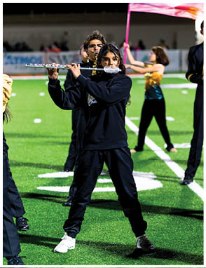 A person wearing a black outfit is playing a flute on a sports field with other people in the background, including one holding a pink flag. Description generated by AI