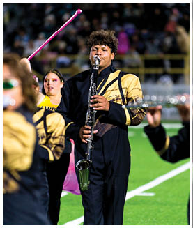 A person in a black and gold marching band uniform is playing a clarinet on a field with other band members and a crowd in the background. Description generated by AI