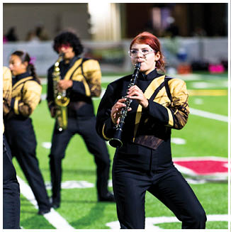 A person in a black and gold marching band uniform is playing a clarinet on a football field with other band members in the background. Description generated by AI