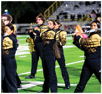 Adults in matching black and gold uniforms are playing flutes and a saxophone on a football field during a nighttime performance. Description generated by AI