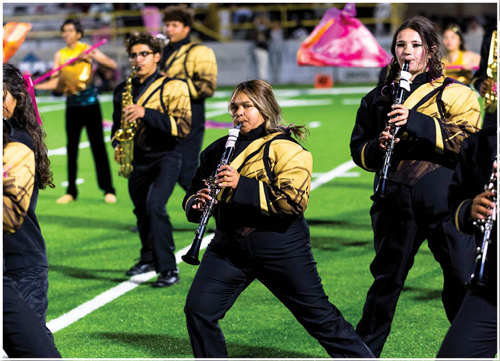 People in matching uniforms are playing musical instruments on a field with a crowd seated in the background. Description generated by AI