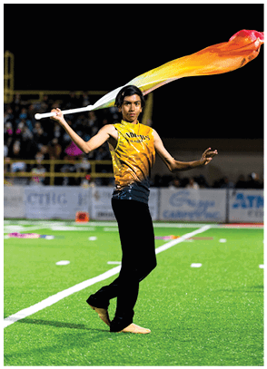 A person wearing a yellow and black sleeveless top and black pants is performing a flag spinning routine on a green sports field at night. Description generated by AI
