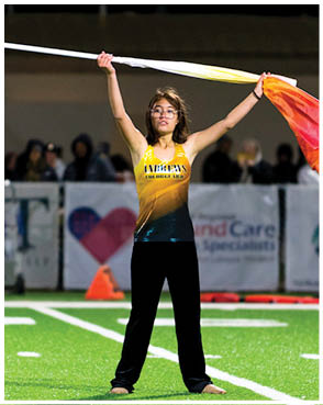 A person wearing a yellow sports uniform is holding a flag with red and orange colors raised above their head at an outdoor event with a dark background. Description generated by AI