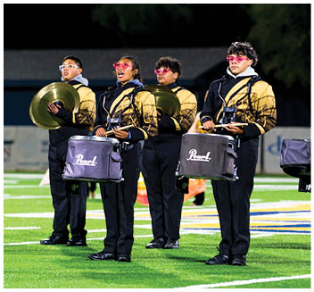 Four people in matching band uniforms stand on a field at night holding percussion instruments and cymbals, wearing pink-tinted glasses. Description generated by AI