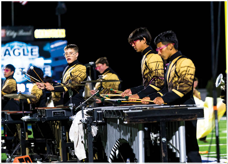 Adults wearing matching band uniforms are playing percussion instruments on a football field at night. Description generated by AI