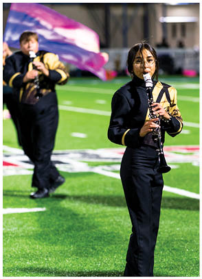 A person wearing glasses and a black and gold uniform is playing a clarinet on a sports field with blurred background elements. Description generated by AI