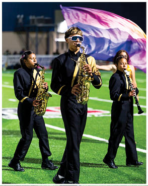 People in marching band uniforms are playing saxophones and a clarinet on a football field with a colorful flag waving in the background. Description generated by AI