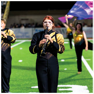 A person with red hair and glasses is playing a clarinet while wearing a black and yellow marching band uniform on a field with blurred people and flags in the background. Description generated by AI