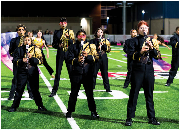People in black and gold marching band uniforms are playing clarinets and saxophones while standing on a football field at night. Description generated by AI