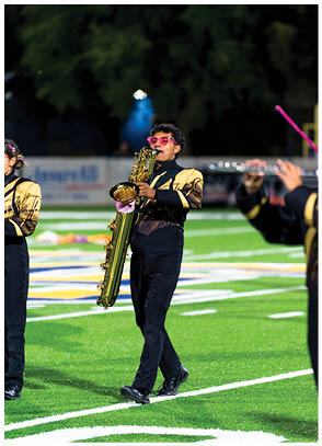 A person wearing a black and gold marching band uniform and pink sunglasses is playing a large saxophone while walking on a football field. Description generated by AI