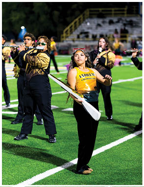 A person in a gold and black uniform holding a white rifle prop stands on a football field with other people playing flutes in the background. Description generated by AI