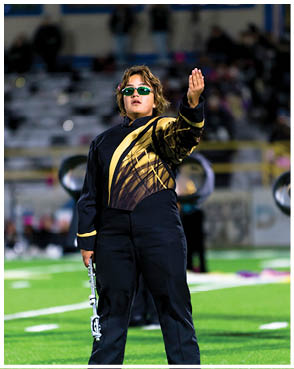A person wearing a black and gold marching band uniform and sunglasses stands on a football field holding a flute and raising one hand. Description generated by AI