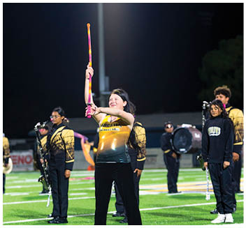 A person in a yellow and black uniform is holding a colorful baton upright on a sports field at night while other people in similar uniforms stand in the background with musical instruments. Description generated by AI