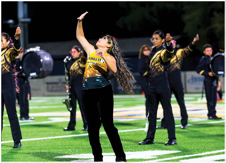 A group of people in matching black and gold uniforms perform a coordinated routine on a sports field at night, with one person in the foreground raising an arm and looking upward. Description generated by AI