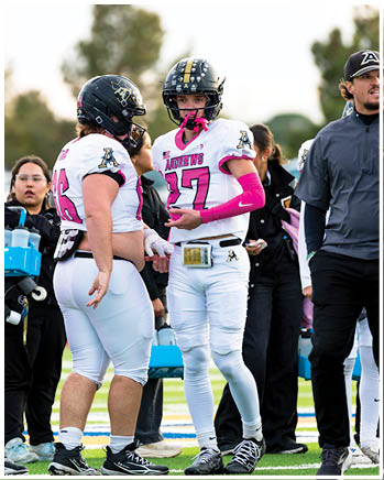 Two people wearing football helmets and white jerseys with pink accents are engaged in conversation on a field. Description generated by AI