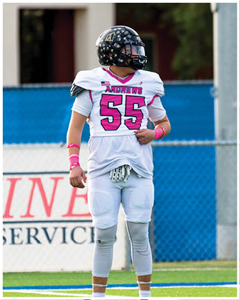 A person wearing a white football jersey with the number 55 and the name Andrews, along with a black helmet, stands on a field with a blue fence and blurred background. Description generated by AI