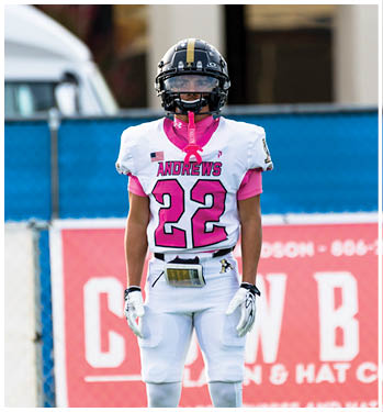 A person wearing a football helmet and a white jersey with pink accents stands outdoors with a blurred background. Description generated by AI