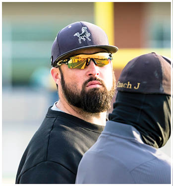A person wearing a dark cap with a horse logo, reflective sunglasses, and a black shirt is looking slightly upward while standing next to another person wearing a dark cap with partially visible text. Description generated by AI