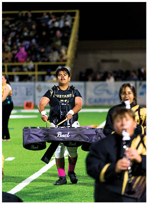A person wearing a black shirt with “Mustangs" on it is playing a marching percussion instrument on a football field during a nighttime event with other musicians and a crowd in the background. Description generated by AI
