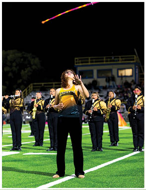 A person wearing glasses and a yellow sports jersey is reaching up as a colorful baton is thrown into the air during a nighttime outdoor event with other people in the background. Description generated by AI