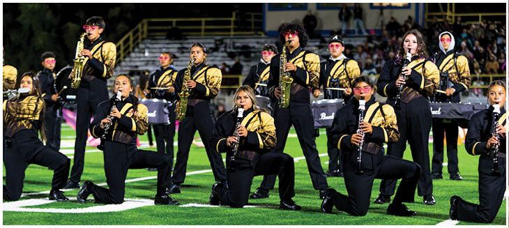 A group of people dressed in matching black and gold uniforms are playing clarinets and saxophones while kneeling and standing on a football field during a nighttime performance. Description generated by AI