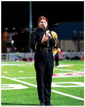 A person wearing a black and yellow marching band uniform is standing on a football field at night playing a clarinet. Description generated by AI