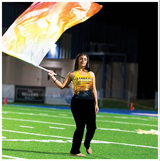 A person wearing a yellow and black outfit is standing on a sports field at night while waving a large flag with red, orange, and white colors. Description generated by AI