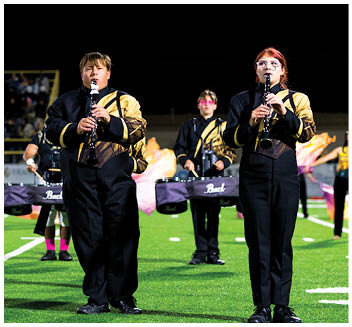 People in matching band uniforms are playing clarinets and drums while standing on a football field during a nighttime event. Description generated by AI