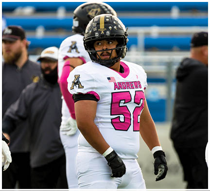 A person wearing a white and pink football uniform with the number 52 stands on a field with other people in the background. Description generated by AI