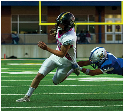 One person in a blue football uniform is diving to tackle another person in a white football uniform who is running with the ball on a green field. Description generated by AI