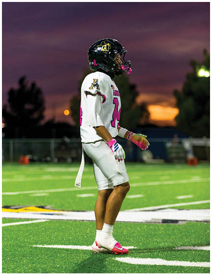 A person wearing a football uniform with a black helmet and white and pink gear stands on a green field at sunset. Description generated by AI