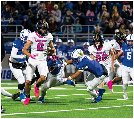 A group of football players in blue and white uniforms are actively trying to tackle a player in a white and pink uniform running with the ball on a brightly lit field with a referee observing nearby. Description generated by AI
