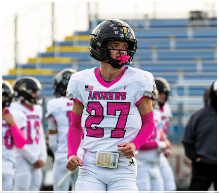 A person wearing a white football uniform with pink accents and a black helmet stands on a field with other players and a coach in the background. Description generated by AI