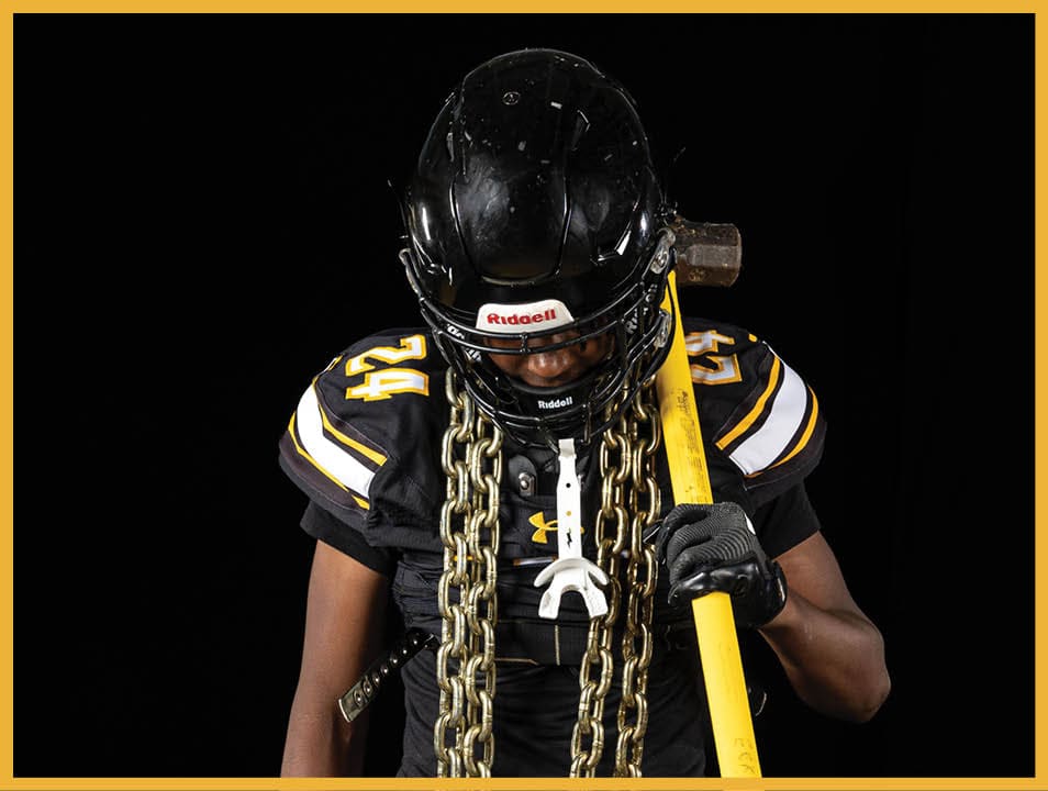 A person wearing a black football uniform and helmet stands holding a yellow sledgehammer with heavy chains draped around their neck against a black background. Description generated by AI