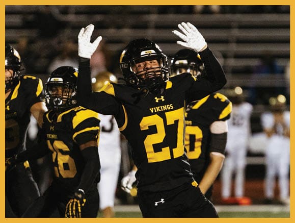 A person wearing a black and yellow Vikings football uniform with the number 27 raises both hands while other players stand nearby on a football field. Description generated by AI