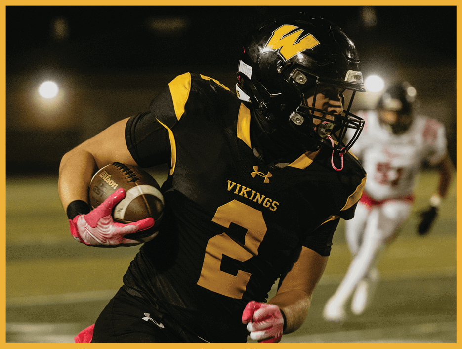 A person wearing a black and yellow Vikings football uniform with the number 2 is running with a football while another player in white follows behind on a lit football field at night. Description generated by AI