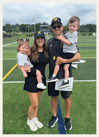 Two adults wearing matching hats and athletic clothing stand on a sports field holding two children who are smiling and dressed in casual outfits. Description generated by AI