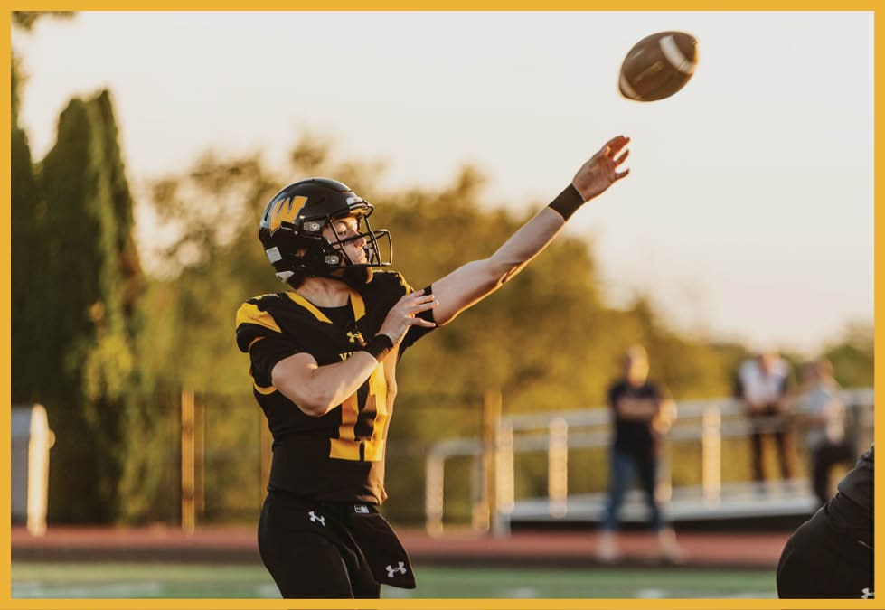 A person wearing a black and yellow football uniform is throwing a football on a field with blurred spectators in the background. Description generated by AI