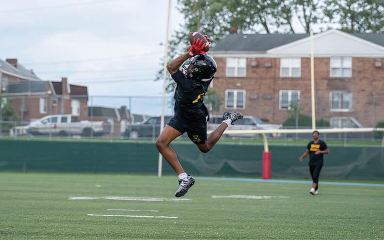 A person wearing a black sports uniform and red gloves is jumping to catch a football on a grassy field with another person standing in the background near a goalpost. Description generated by AI