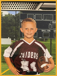 A child with short hair wearing a maroon and white sports jersey is standing outdoors in front of a green field and trees. Description generated by AI