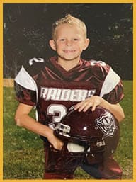A child with short curly hair is smiling and wearing a dark sports jersey with a white number on the shoulder. Description generated by AI